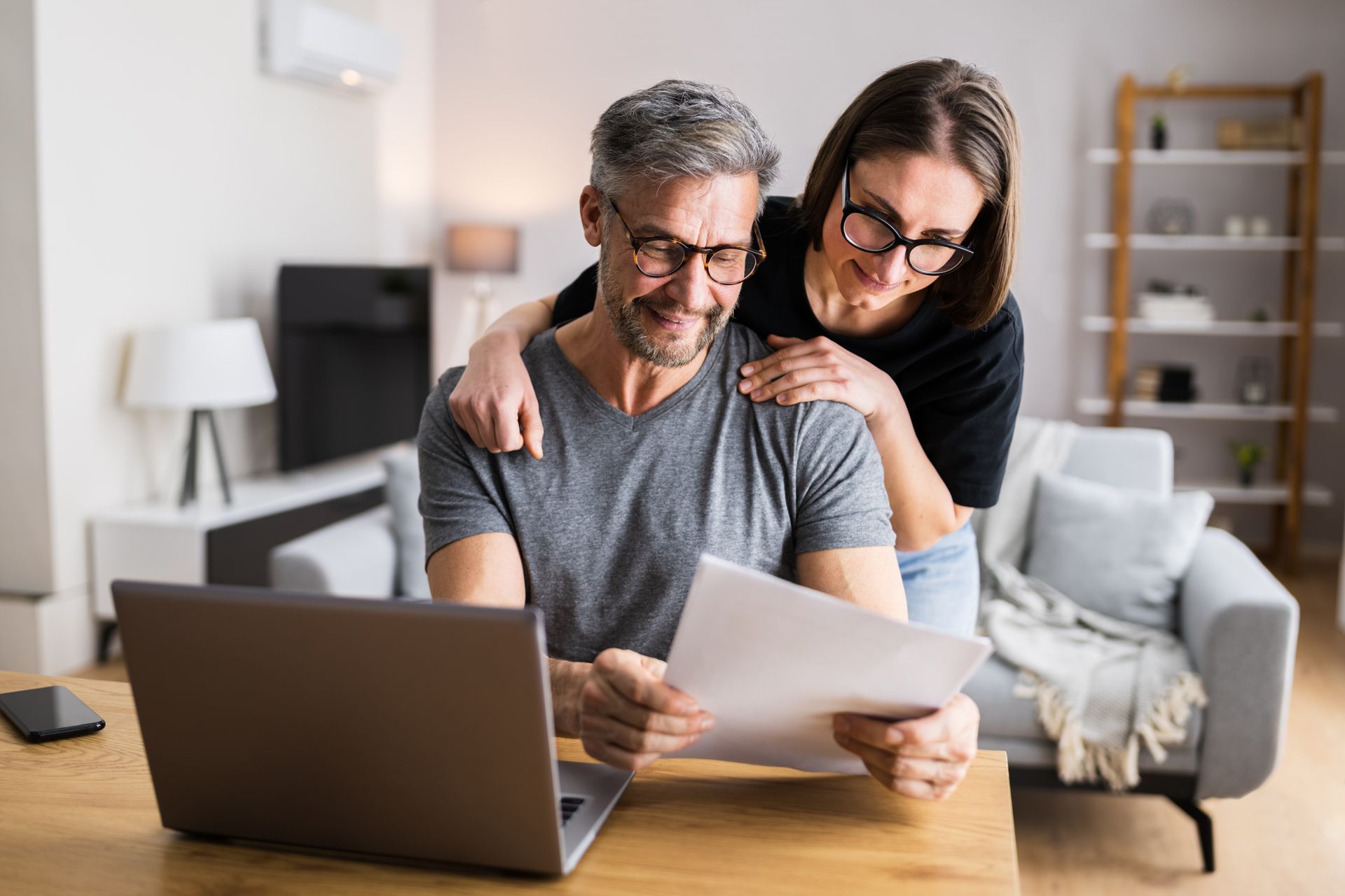 Two people reviewing papers together at a laptop in a bright living room