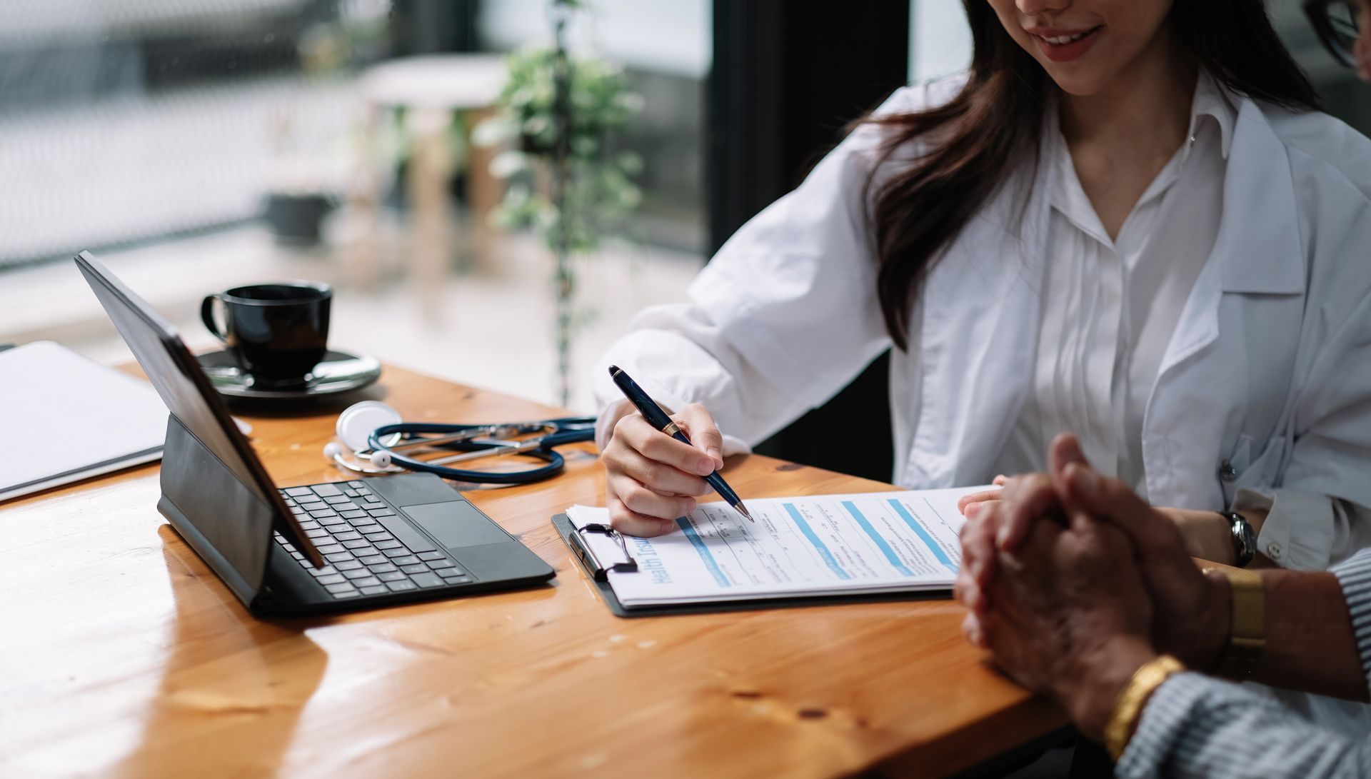 Two people discussing paperwork at a wooden table with a laptop and coffee in a bright office