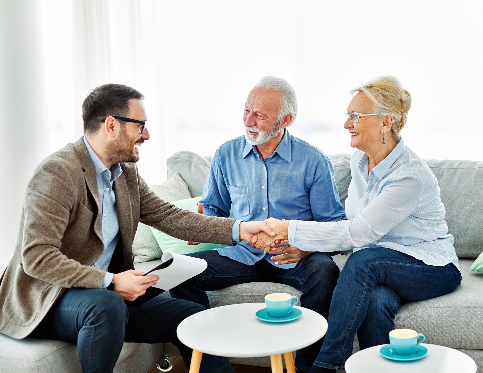 Three people in a bright living room shake hands while seated on a sofa with coffee cups on small tables.