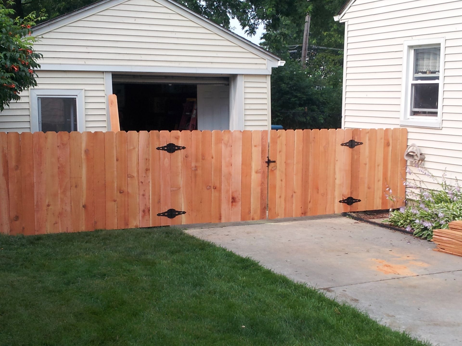 A wooden fence with black hinges and latch encloses a driveway between a garage and a house.