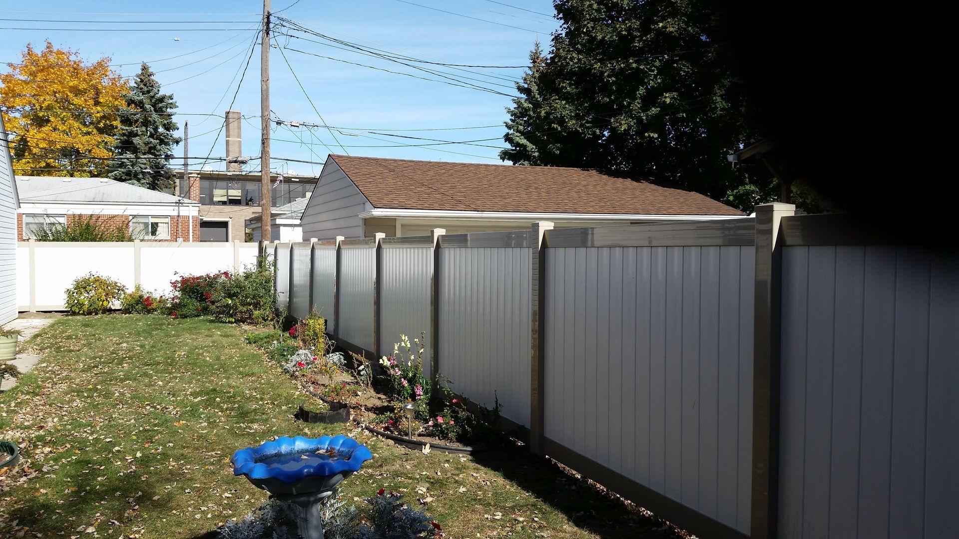 Backyard with white fence, grass, and a small brown-roofed structure; trees and sky in background.