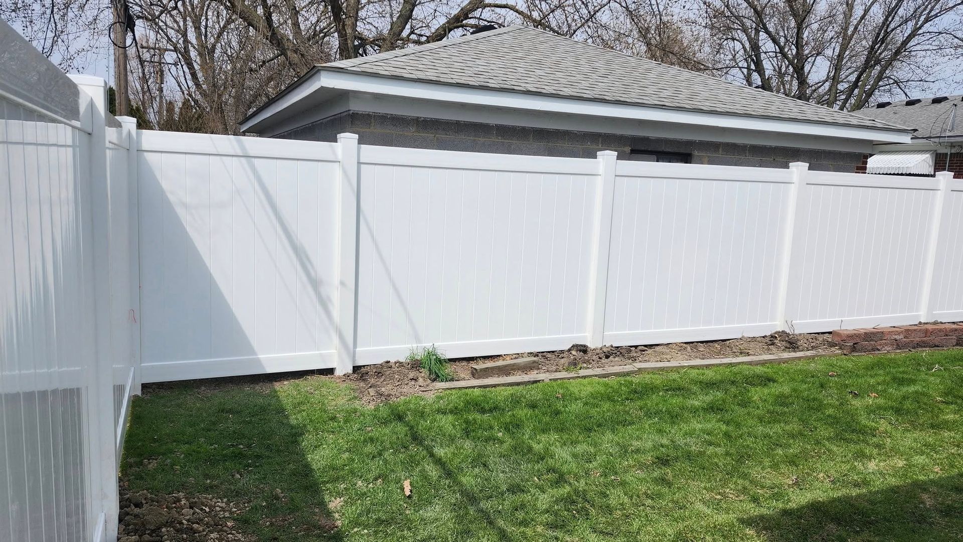 White vinyl fence surrounds a grassy backyard, next to a gray roofed building.