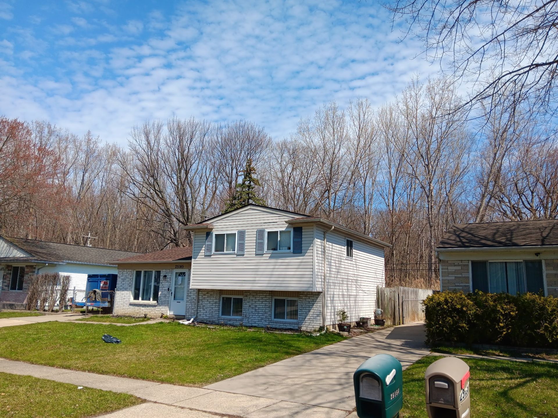 Two-story house with white siding and a driveway, flanked by trees and grass under a blue sky.