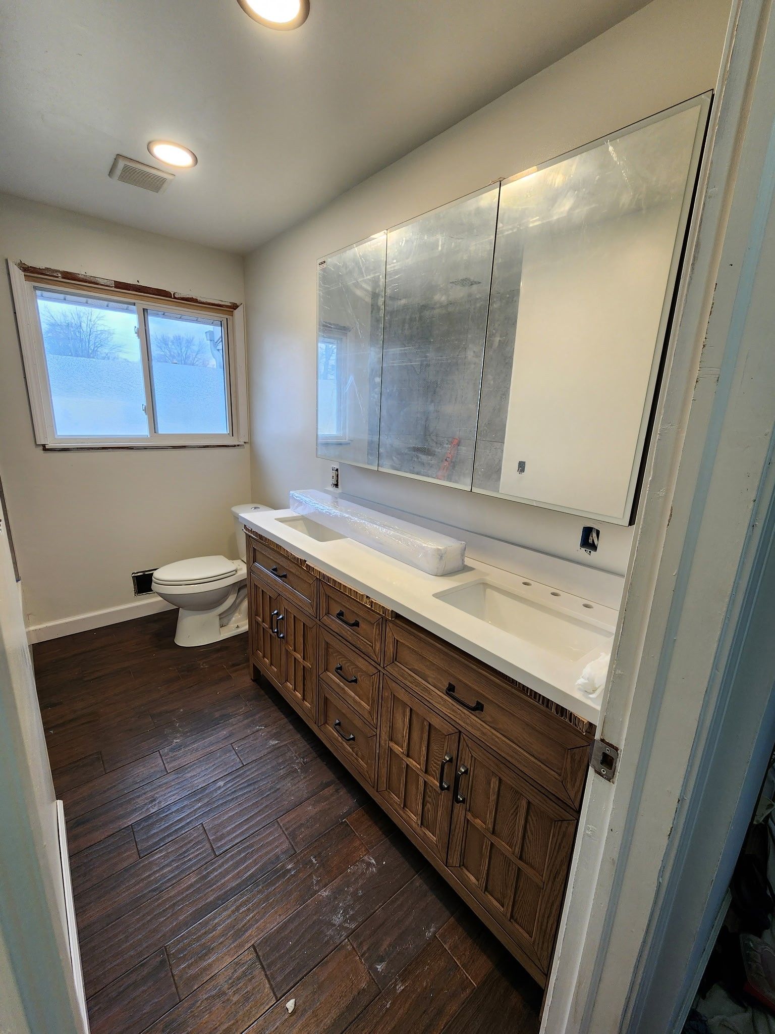 Bathroom with a wooden vanity, white countertop, large mirror, and toilet. Dark brown floor.