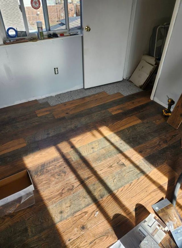 Room with new flooring installation: Wooden planks on the floor with sunlight creating shadows. Door and window visible.
