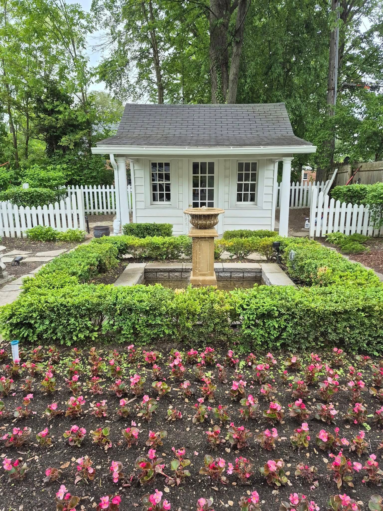 Small white playhouse with picket fence, surrounded by a garden with flowers and a fountain.