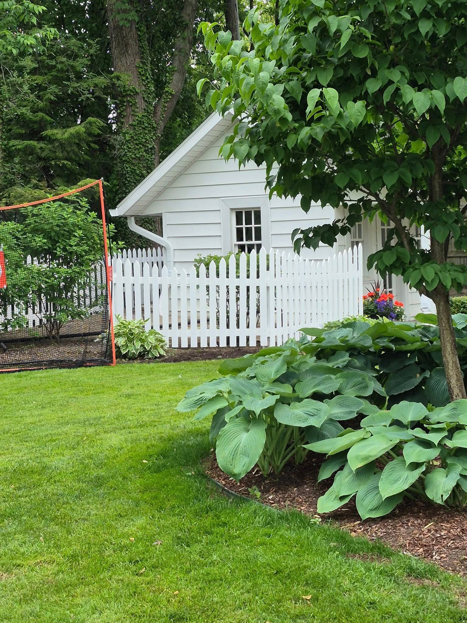 White shed with picket fence, surrounded by green plants and lawn.