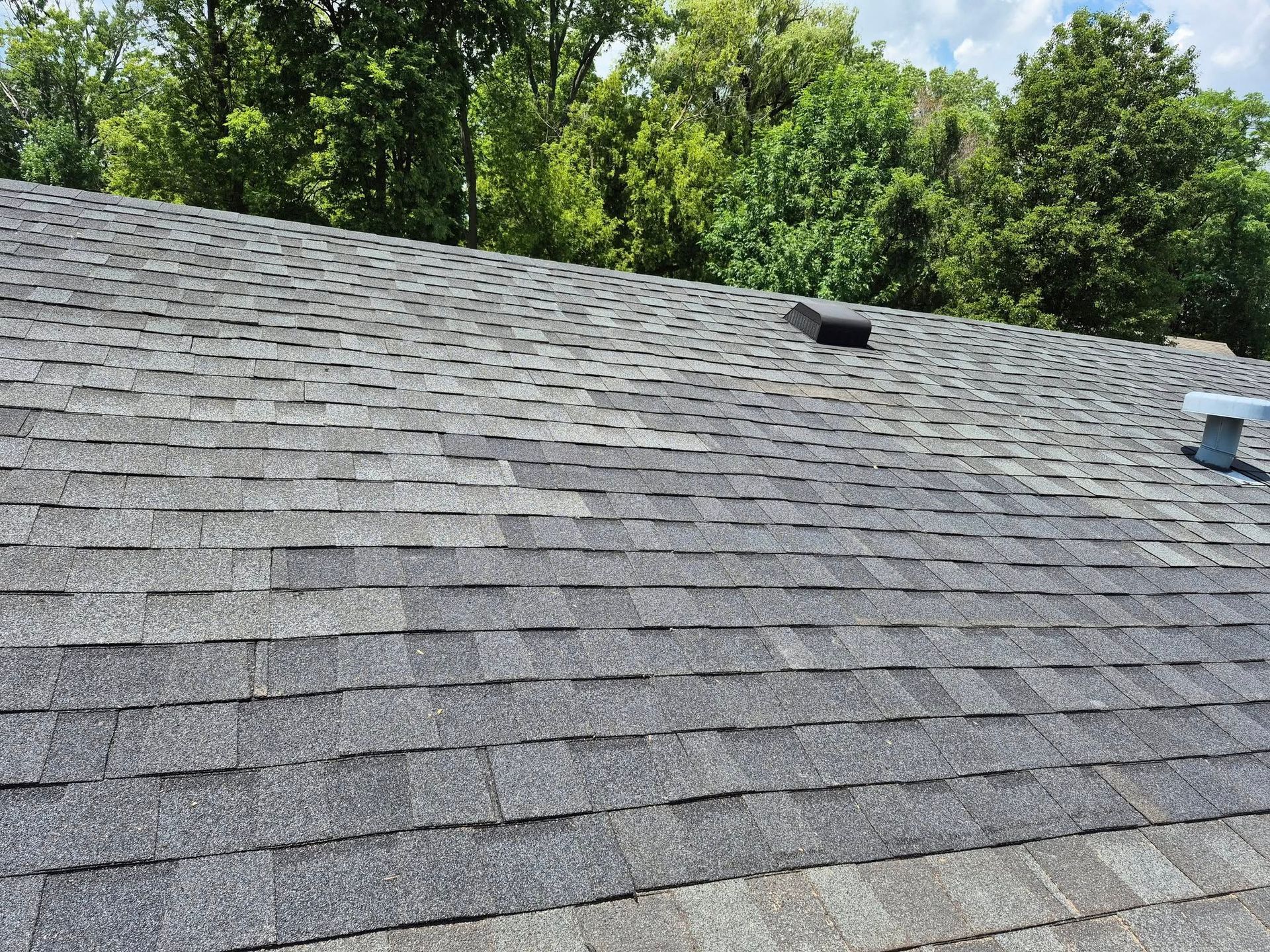 Gray asphalt shingle roof with a black vent against a backdrop of green trees under a cloudy sky.