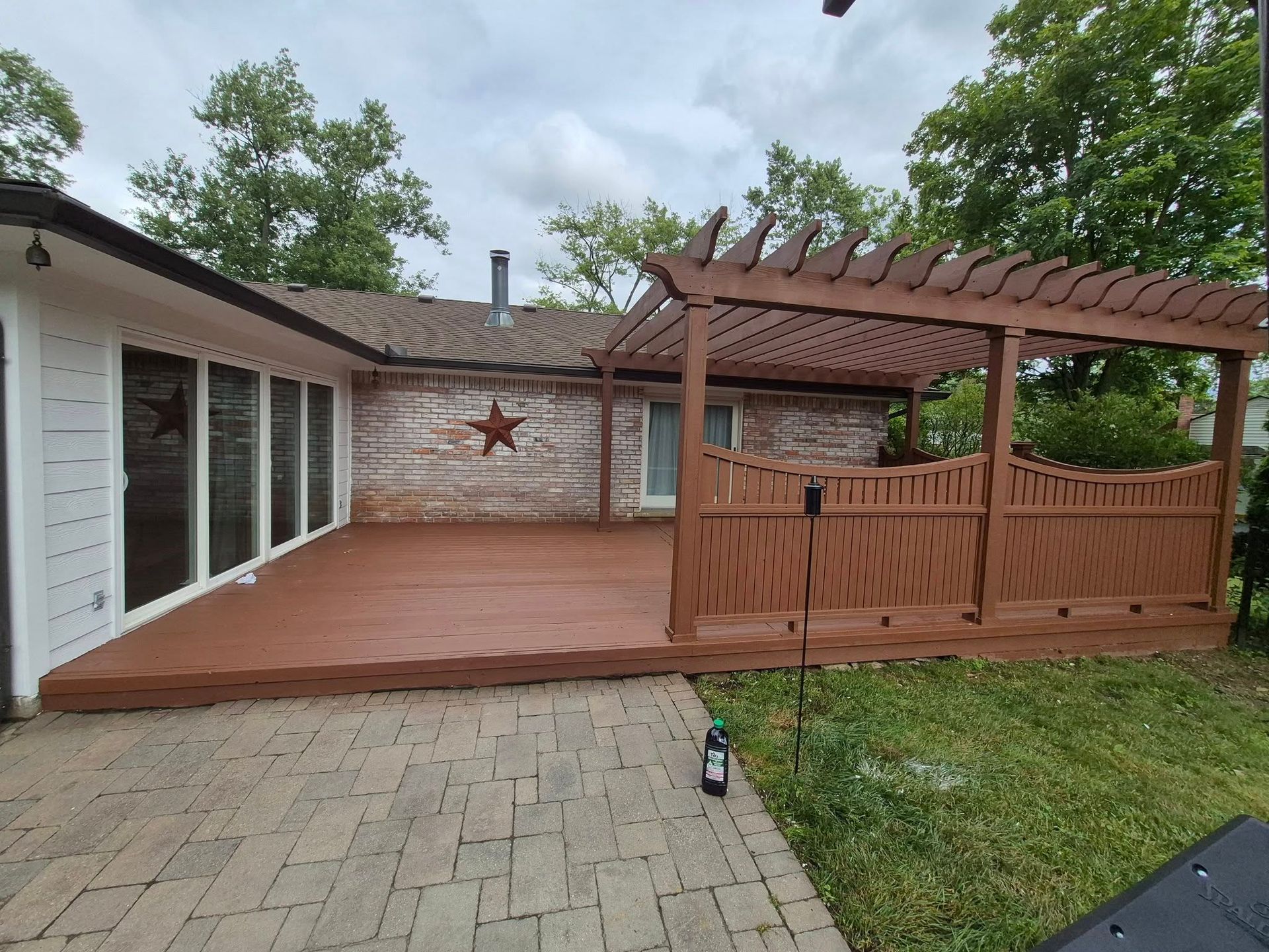 A brown wooden deck with pergola and brick house, paved patio, and green grass under a cloudy sky.