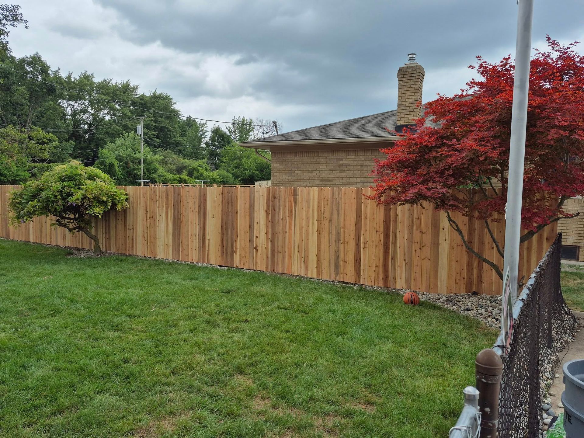 Wooden fence surrounding a grassy yard. House with chimney and red-leafed tree in background. Overcast sky.