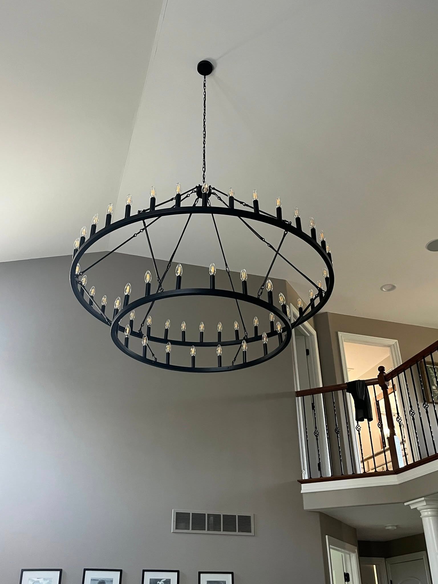 Two-tiered black chandelier hangs from a high ceiling. A staircase with a white railing is in the background.