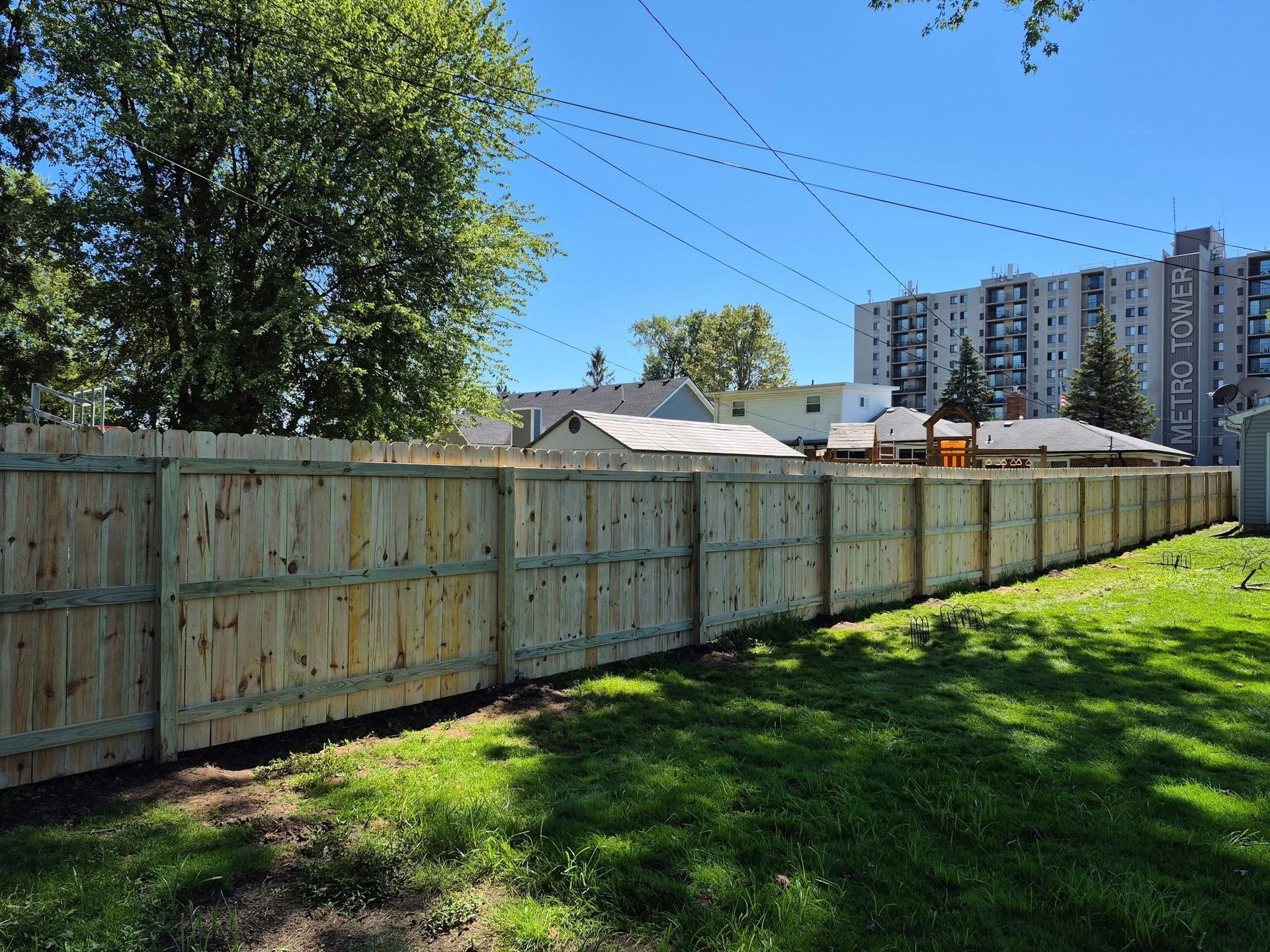 Wooden fence bordering a green lawn, with buildings and trees in the background on a sunny day.