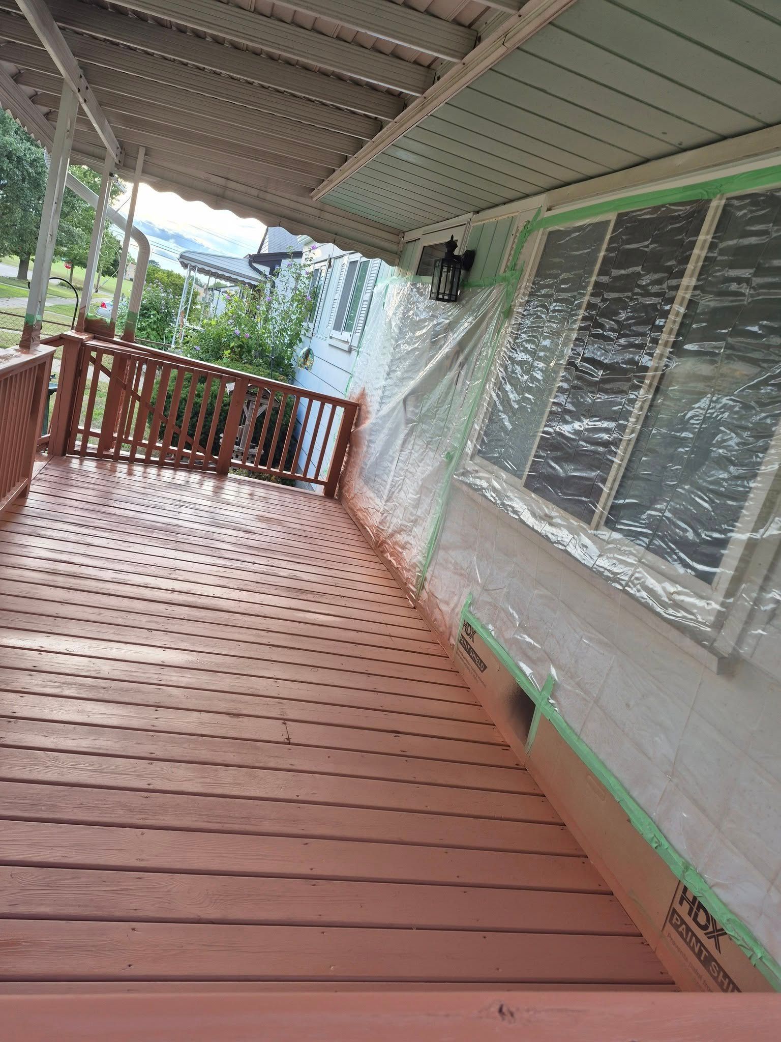 Wooden deck and railing with a house in the background, covered with plastic sheeting for painting.