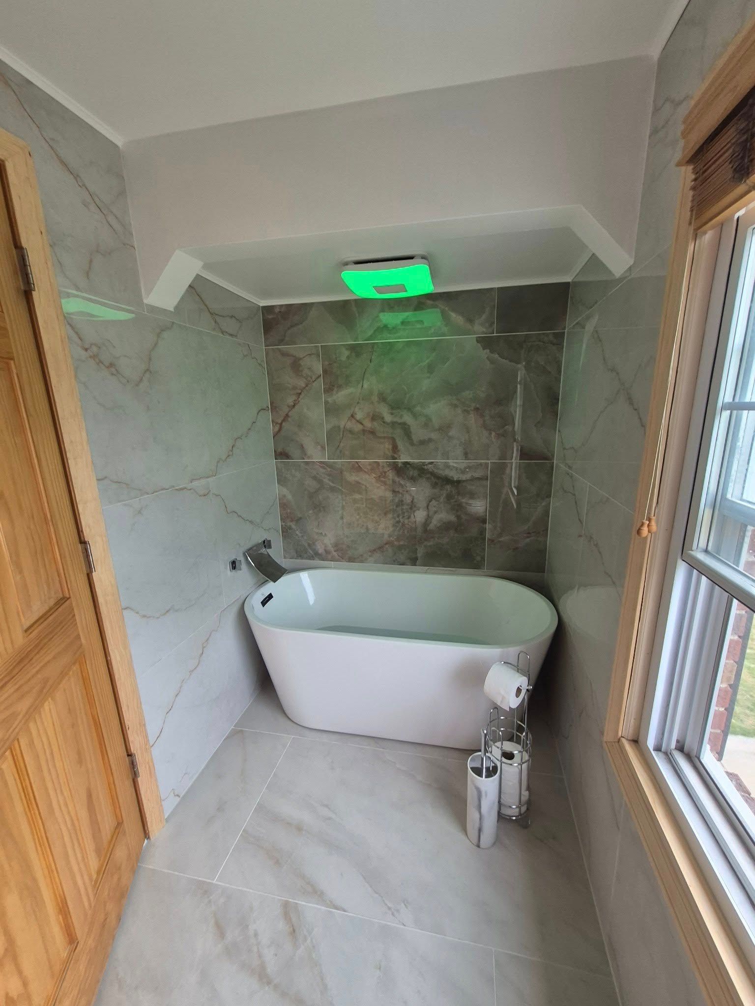 Bathroom with a white tub, marble-look tile, a window, and overhead green light.