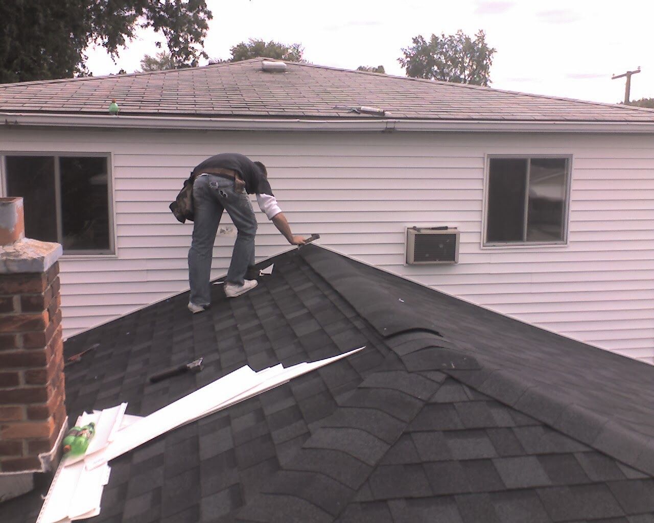 Man on a dark shingle roof repairing shingles near a white house with two windows.