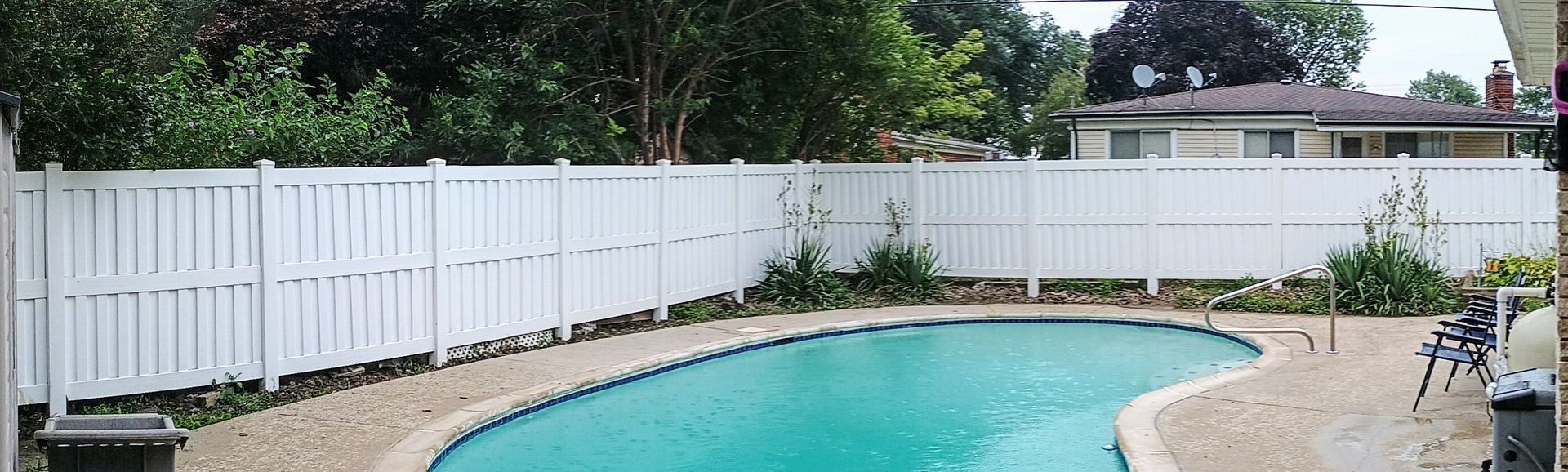 White fence surrounding a blue swimming pool; trees and a house in the background.