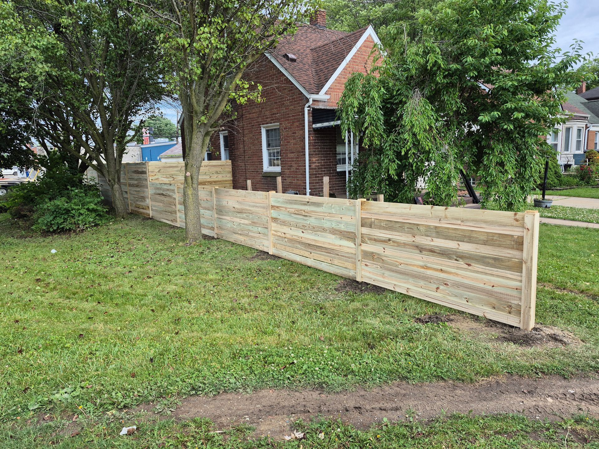 Wooden fence in front yard with a brick house in the background. Green grass, trees, and cloudy sky.