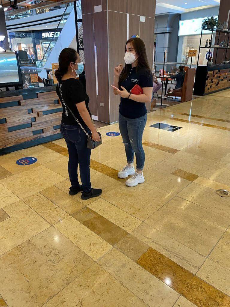 Dos mujeres, una con mascarilla, conversan en un centro comercial. Llevan mascarilla azul, vaqueros y camisa negra.