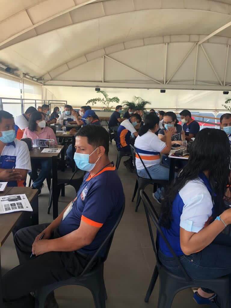 Personas con máscaras sentadas en mesas bajo un techo de dosel, algunas llevan camisas azules y blancas.