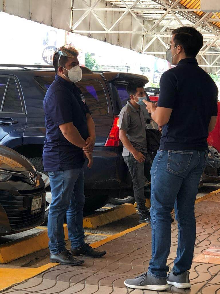 Tres hombres con máscaras hablando cerca de coches estacionados.