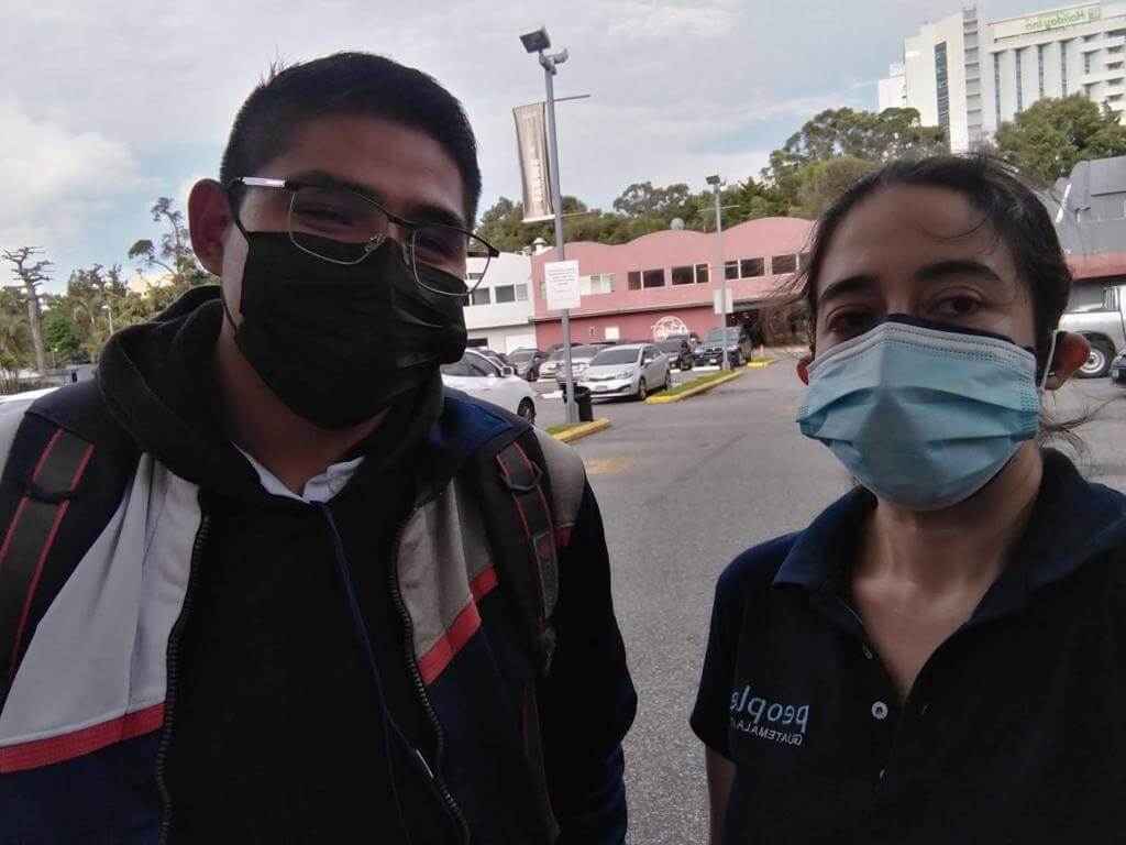 Dos personas con mascarillas afuera de un edificio: un hombre con gafas y una mochila y una mujer con una camisa azul.