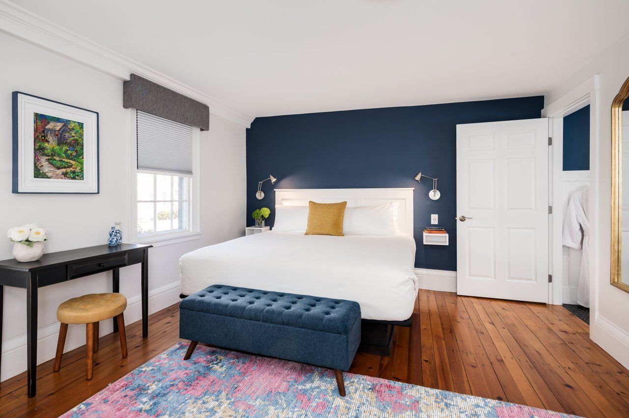 A hotel bedroom featuring a king bed with navy blue accents, a floral rug, and a small writing desk on wood flooring.