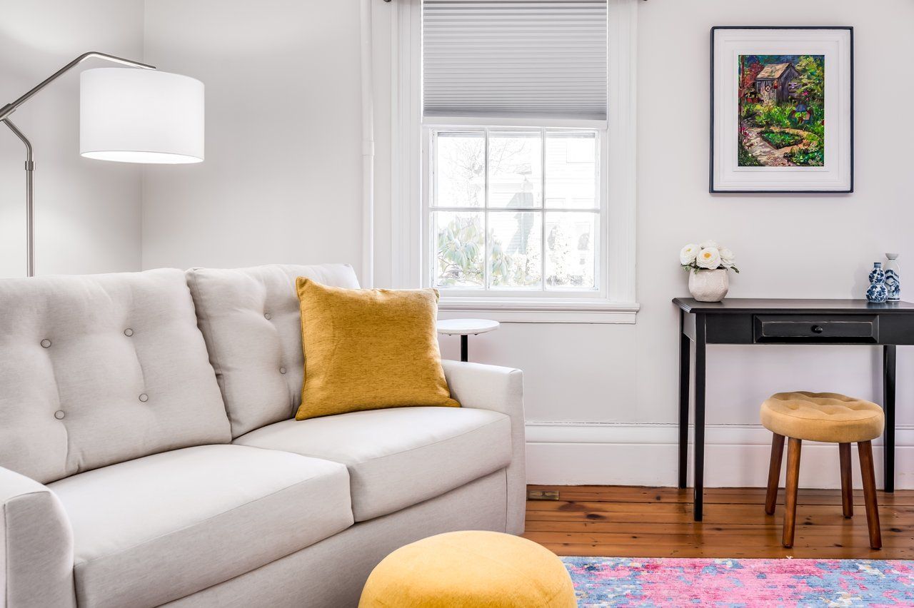 A living room with a light gray sofa, yellow cushion and ottoman, a black desk, and a colorful rug on a wooden floor.