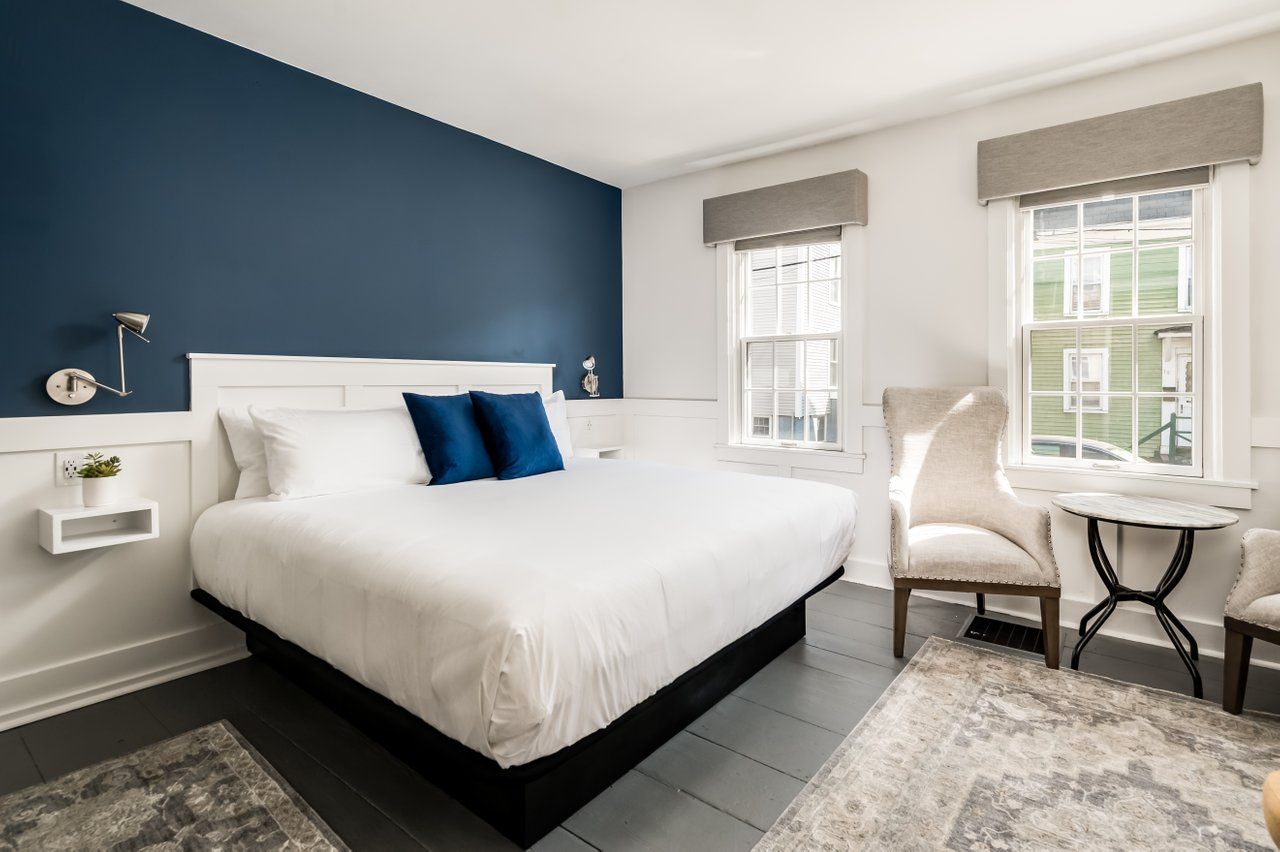 A hotel room with a large bed against a navy wall, white headboard, grey floor, and a small sitting area by the windows.