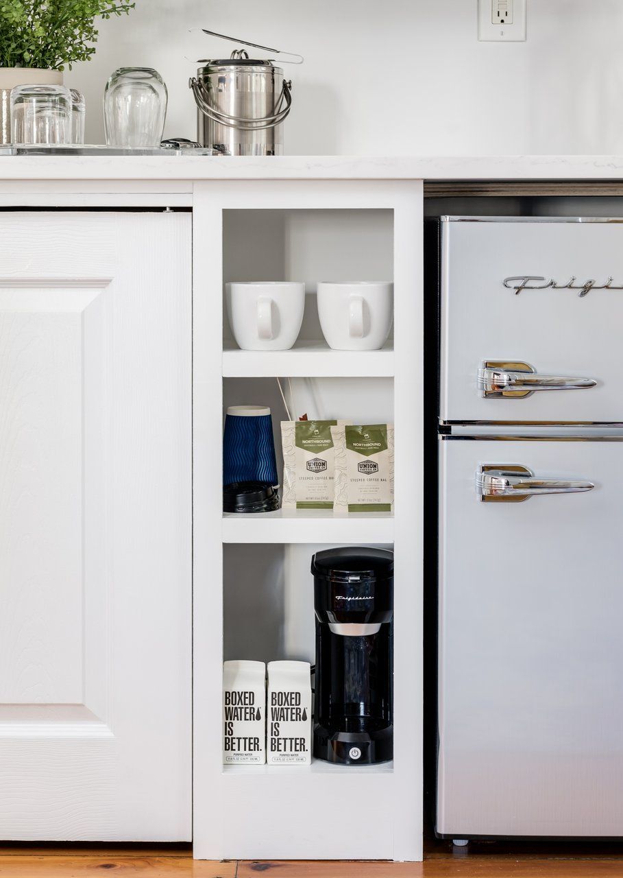 A white narrow kitchen shelf holds coffee mugs, coffee beans, and a brewer, positioned between cabinets and a mini fridge.