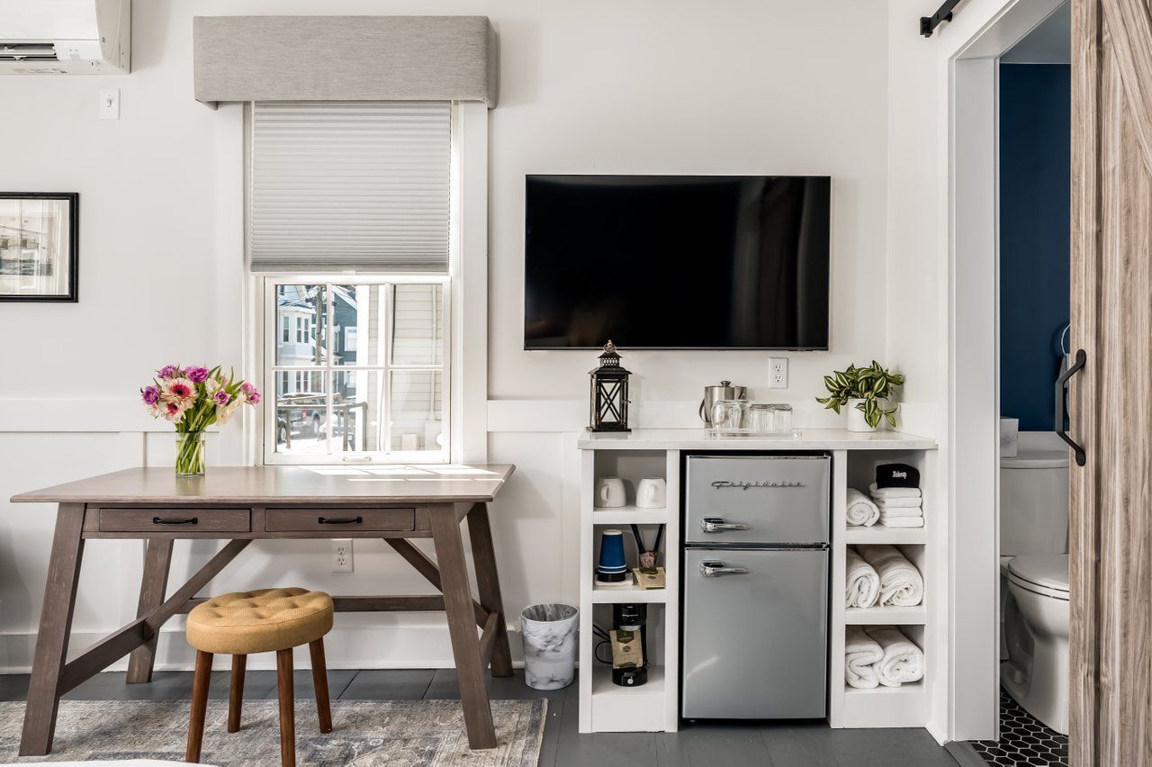 A small wooden table with flowers and a stool next to a TV and white shelving unit holding a fridge and folded towels.