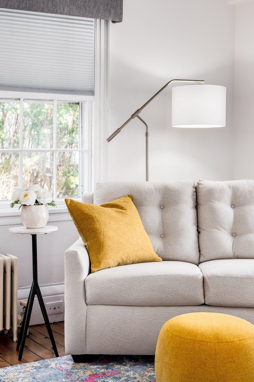 A cozy living room with a beige tufted sofa, a mustard yellow pillow and pouf, a floor lamp, and a vase on a side table.