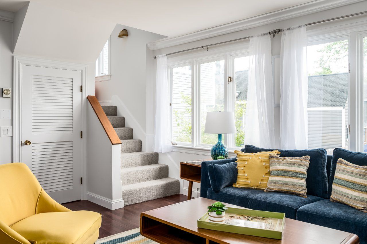 A living room with a navy sofa, yellow armchair, light carpeted stairs, and large windows with white curtains.