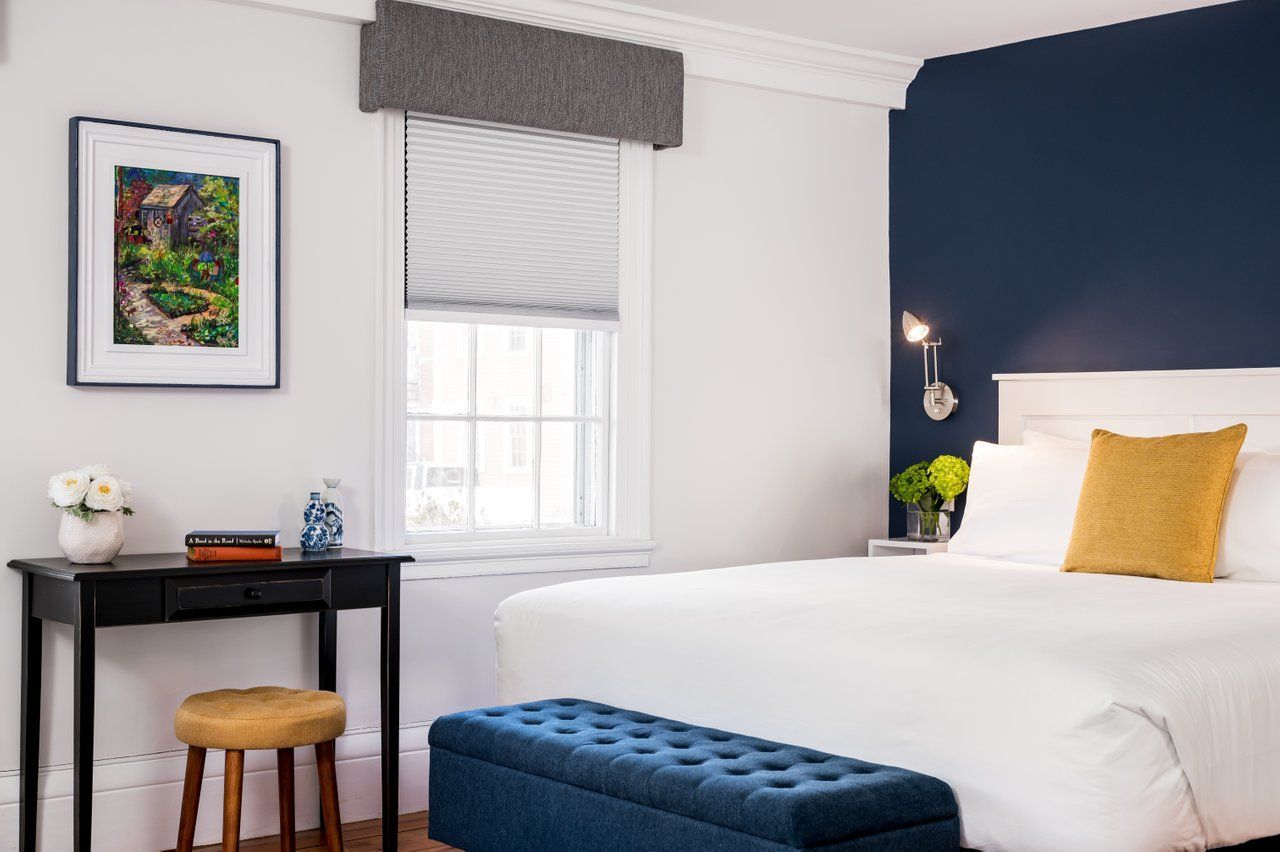 A bedroom featuring a bed with white bedding, a gold pillow, a navy bench, a dark desk, and a wall-mounted reading light.