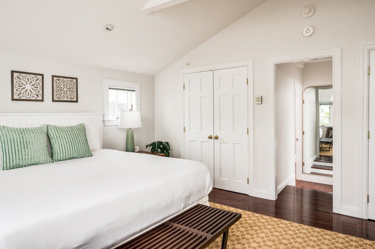 A bedroom with a bed, light green pillows, a bench, a window with a lamp, closed white closet doors, and a doorway.
