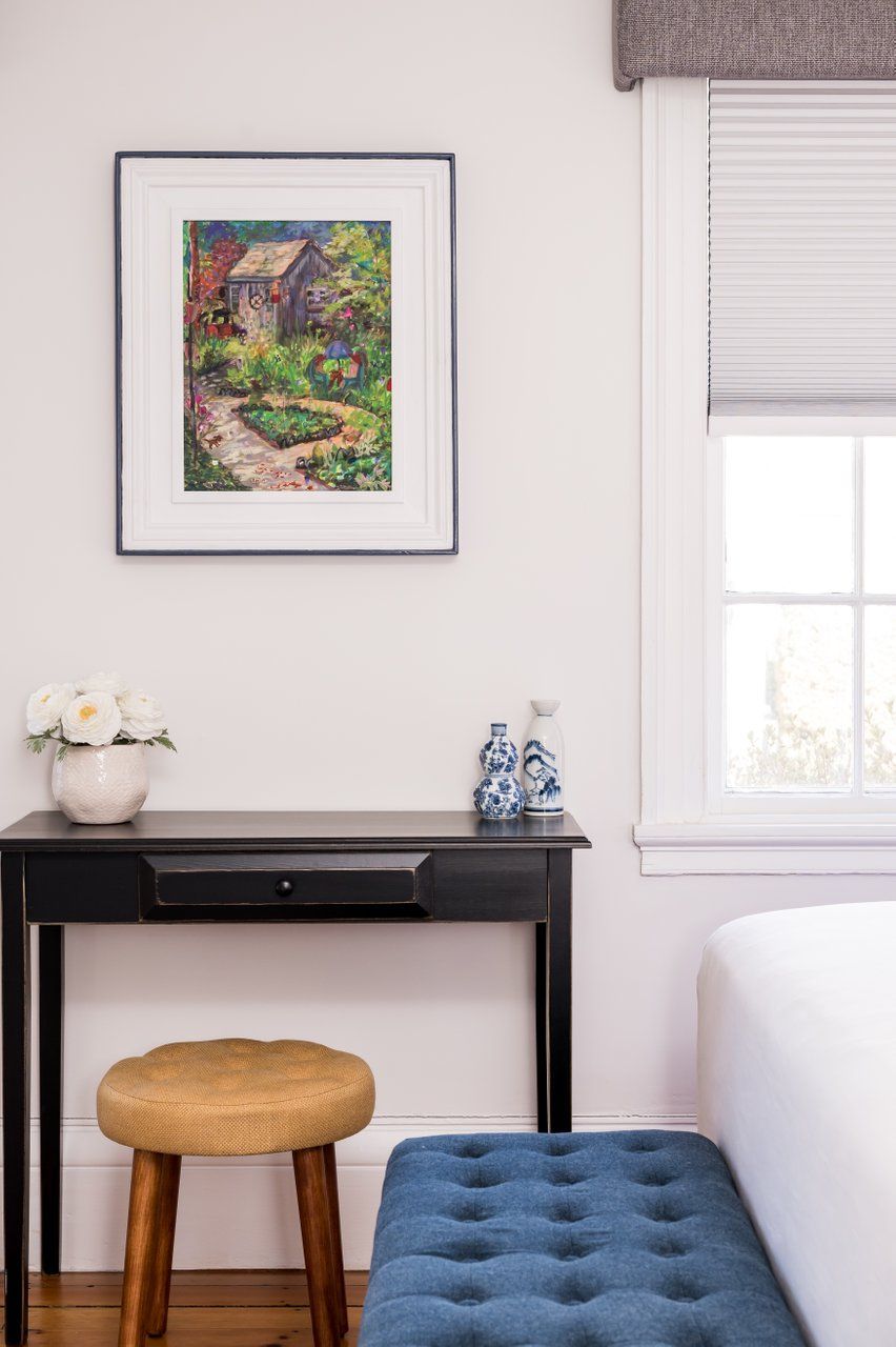 A black desk with a small stool, blue tufted bench, and framed painting on a wall beside a window.