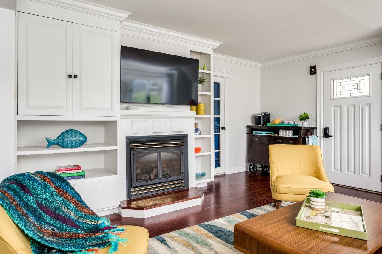 A cozy living room with a fireplace, TV built-ins, yellow armchairs, a patterned rug, and a front door.