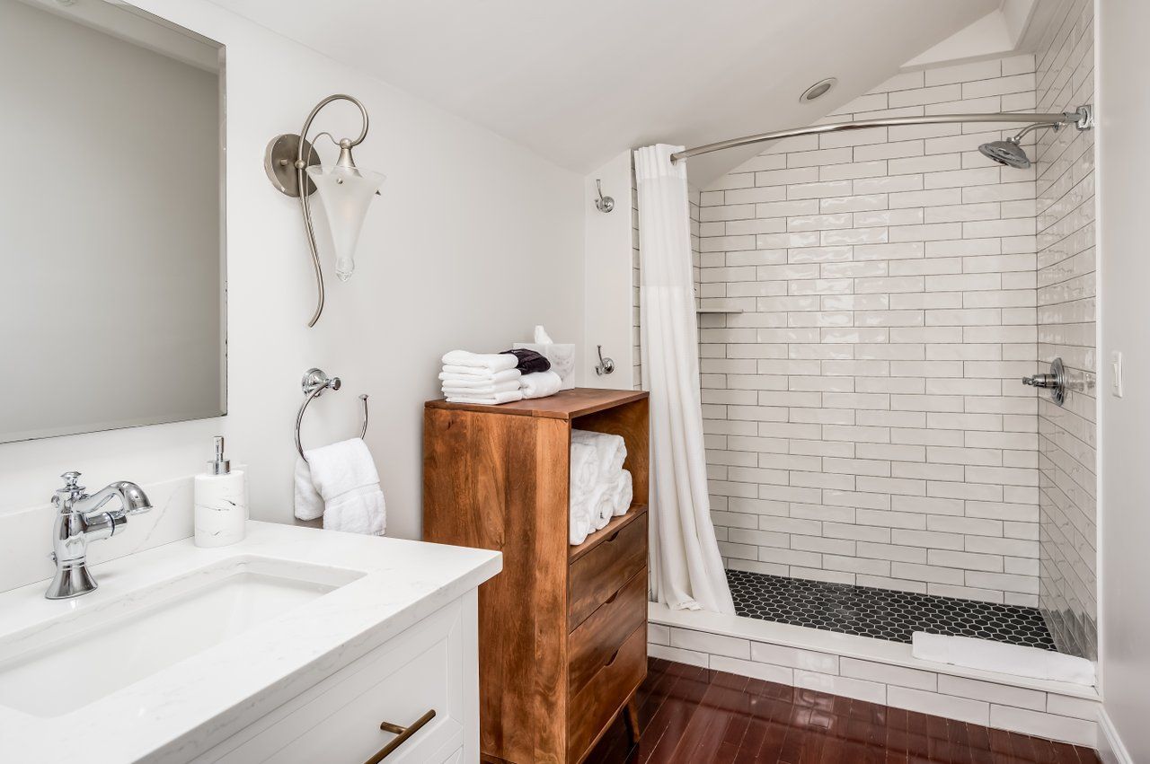 A modern bathroom with a white vanity, a wooden storage unit, a mirror, and a walk-in shower with white tiles.