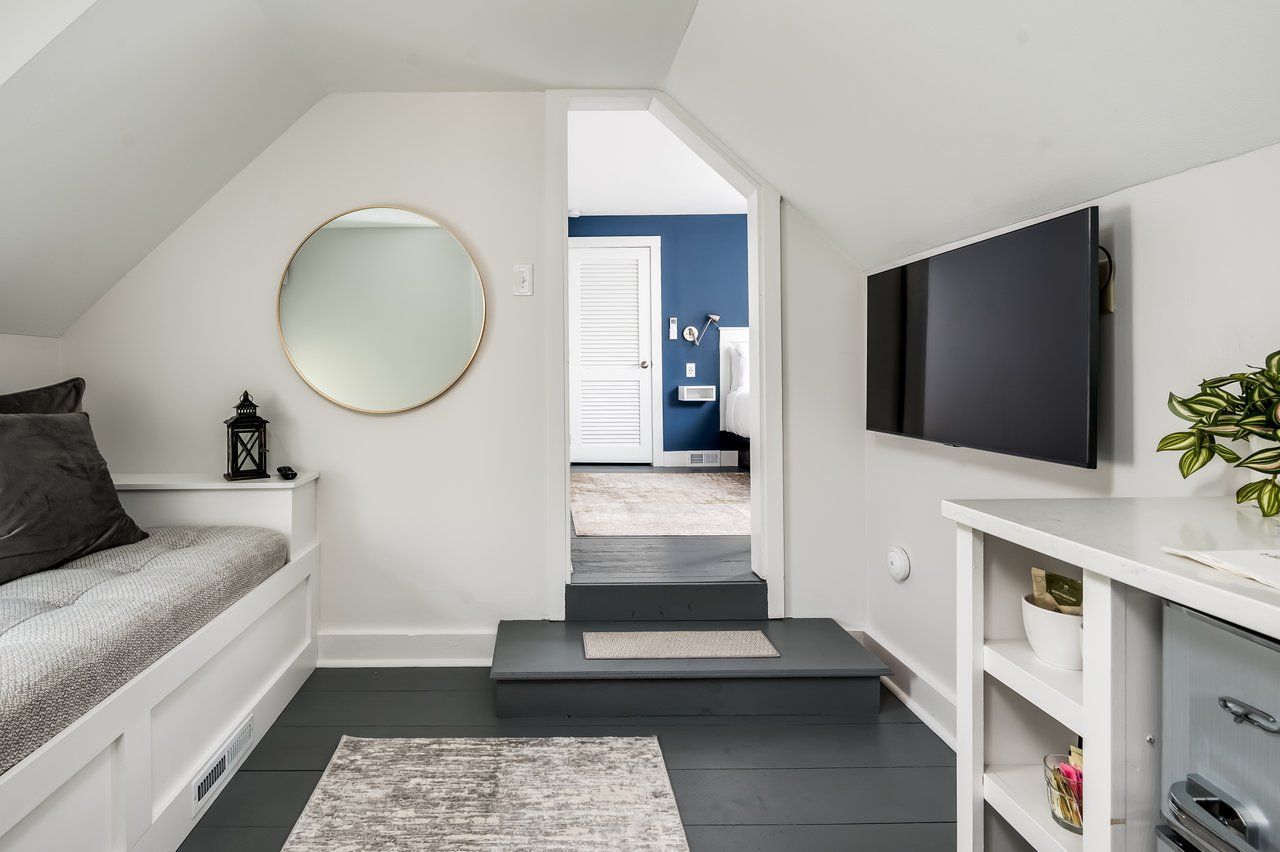 A cozy attic room features a window seat, a round mirror, a wall-mounted TV, and a step leading to an adjacent room.
