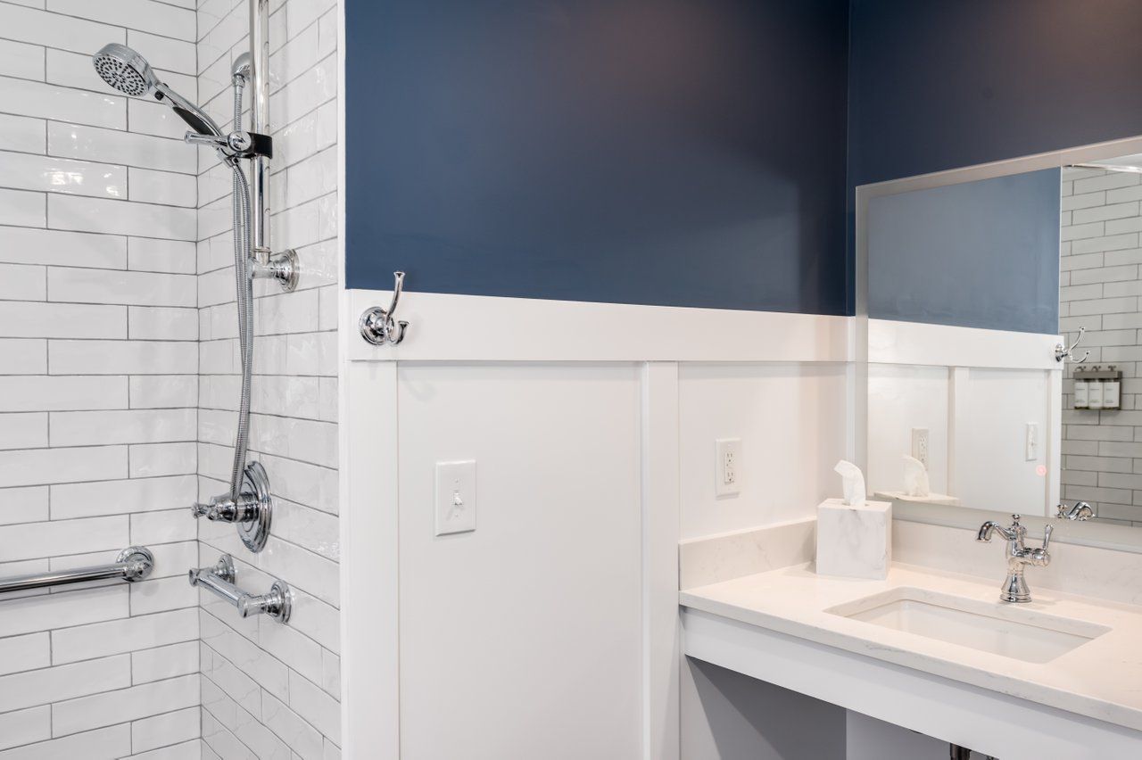 A modern bathroom with a white tiled shower, a navy blue wall, white wainscoting, a vanity, and a mirror.