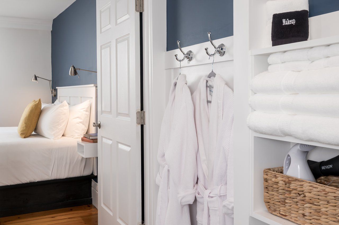 A hotel room interior featuring a bed with white linens, two hanging white robes, and open shelves stacked with towels.