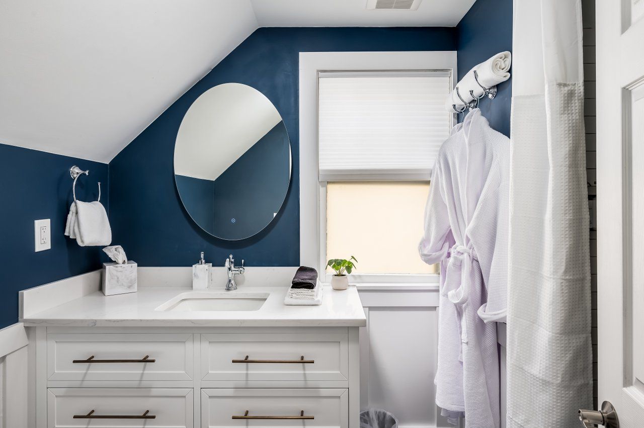 A white vanity with a sink and round mirror against a dark blue wall in a bathroom with a white robe hanging nearby.