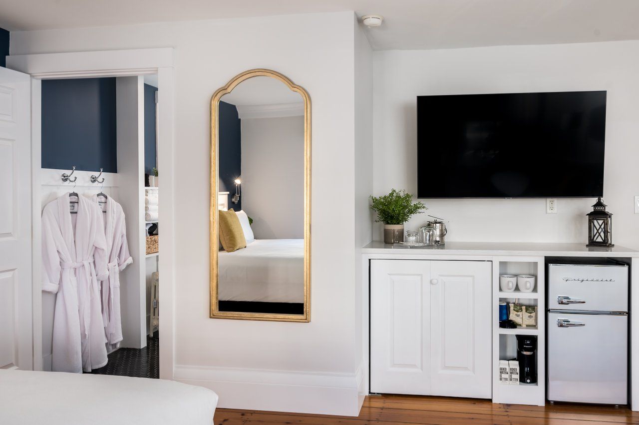 A hotel room scene with white robes hanging in a closet, an ornate gold mirror, a flat-screen TV, and a mini-fridge.