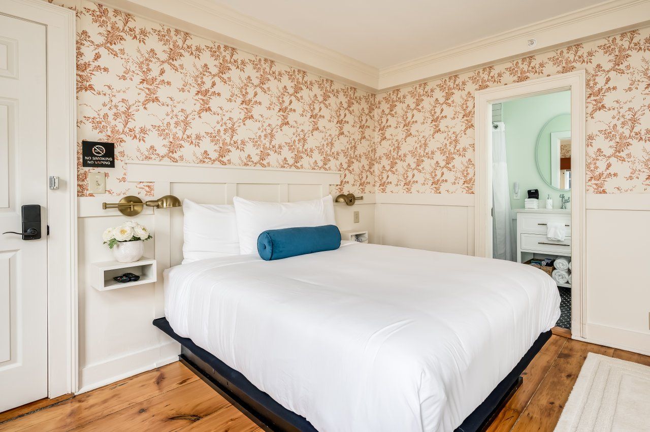 A cozy hotel room featuring floral wallpaper, a white bed with a teal bolster pillow, and a doorway to a bathroom.