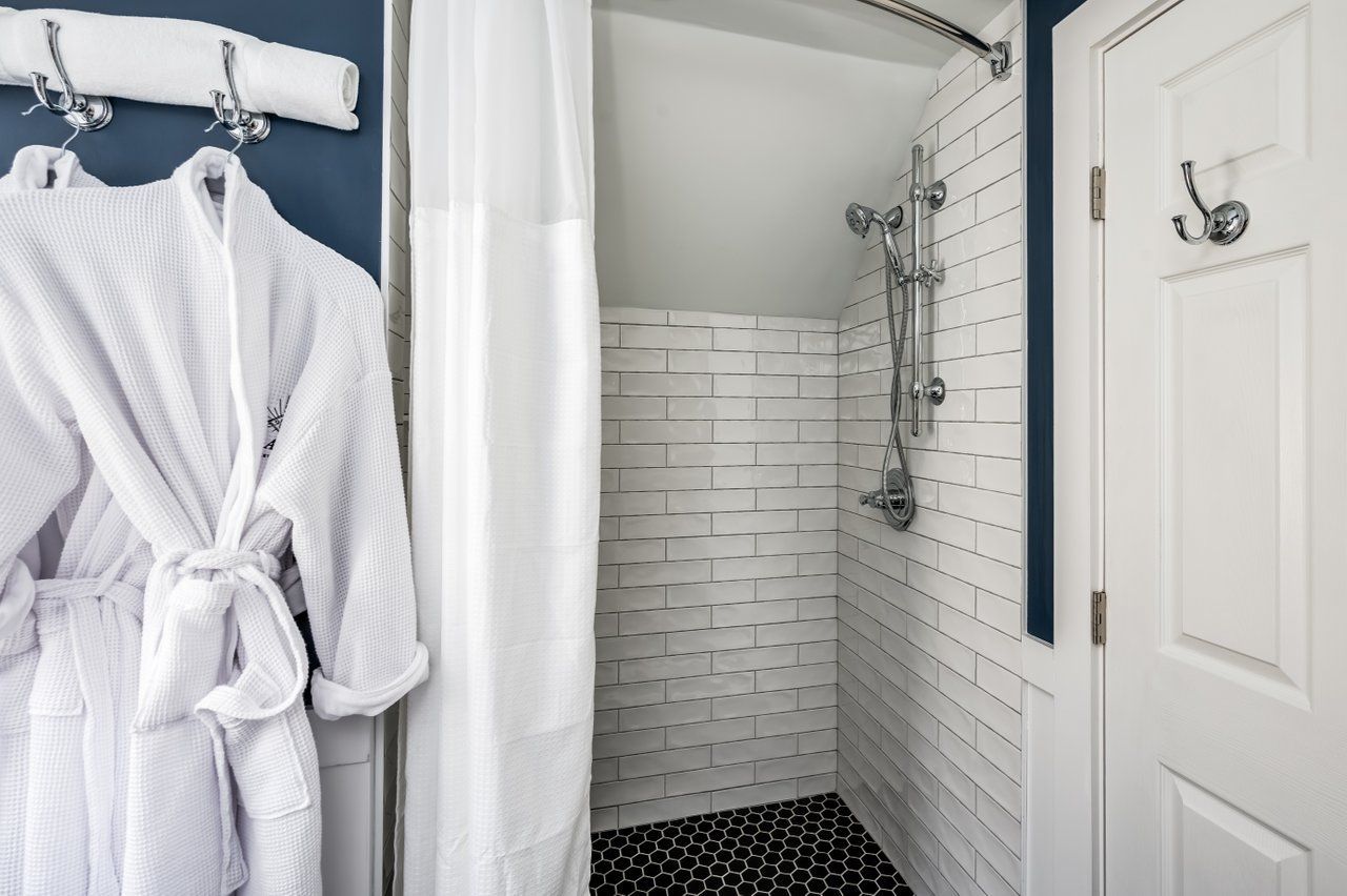 A tiled shower stall next to a door with a robe hanging on a wall hook in a room with dark blue walls.