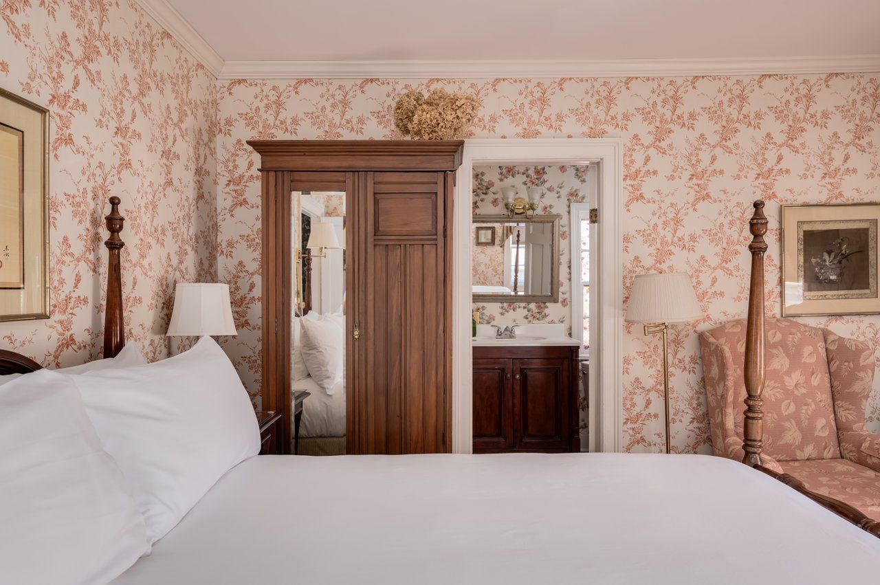 A cozy bedroom featuring floral wallpaper, a wooden wardrobe with a mirror, a bed with white linens, and a patterned chair.