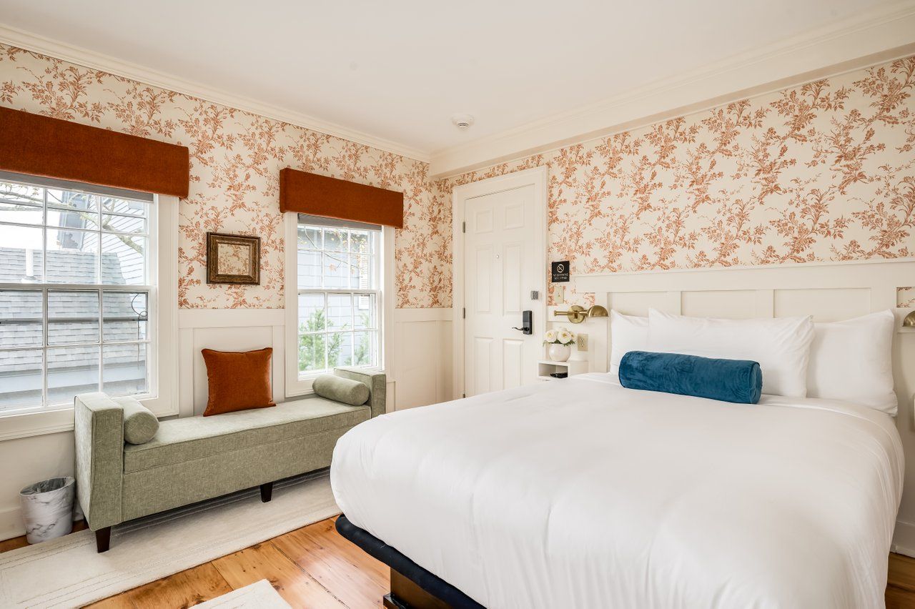 A hotel bedroom with floral wallpaper, a white bed with a blue bolster pillow, and a green bench seat under two windows.