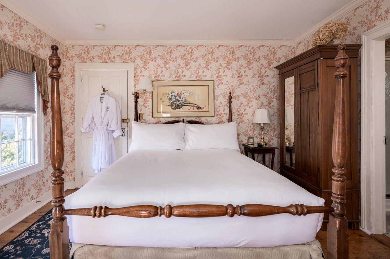 A bedroom with a wooden four-poster bed, floral wallpaper, a wardrobe, and a white robe hanging on the door.