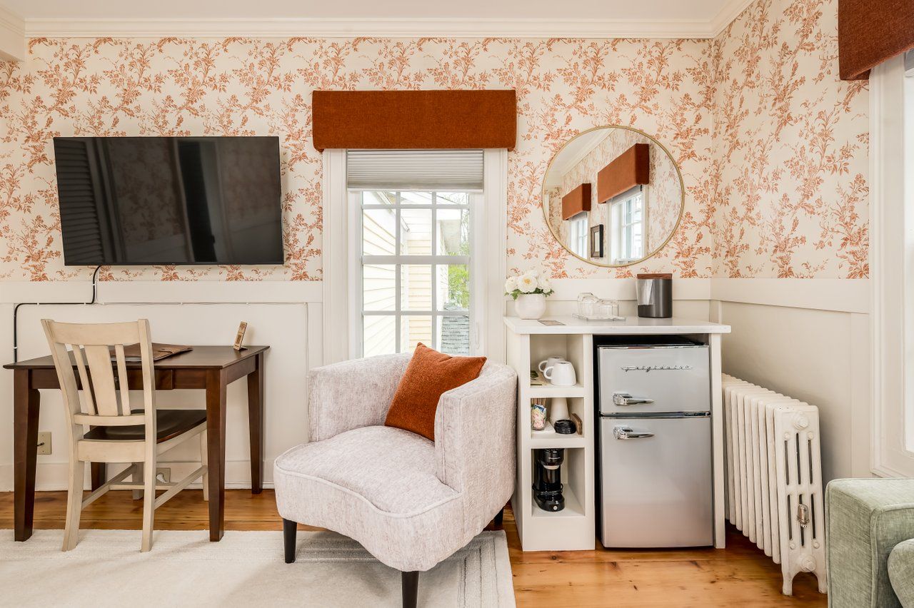A room corner with floral wallpaper, a desk, a TV, a white armchair, a mini-fridge unit, and a radiator.