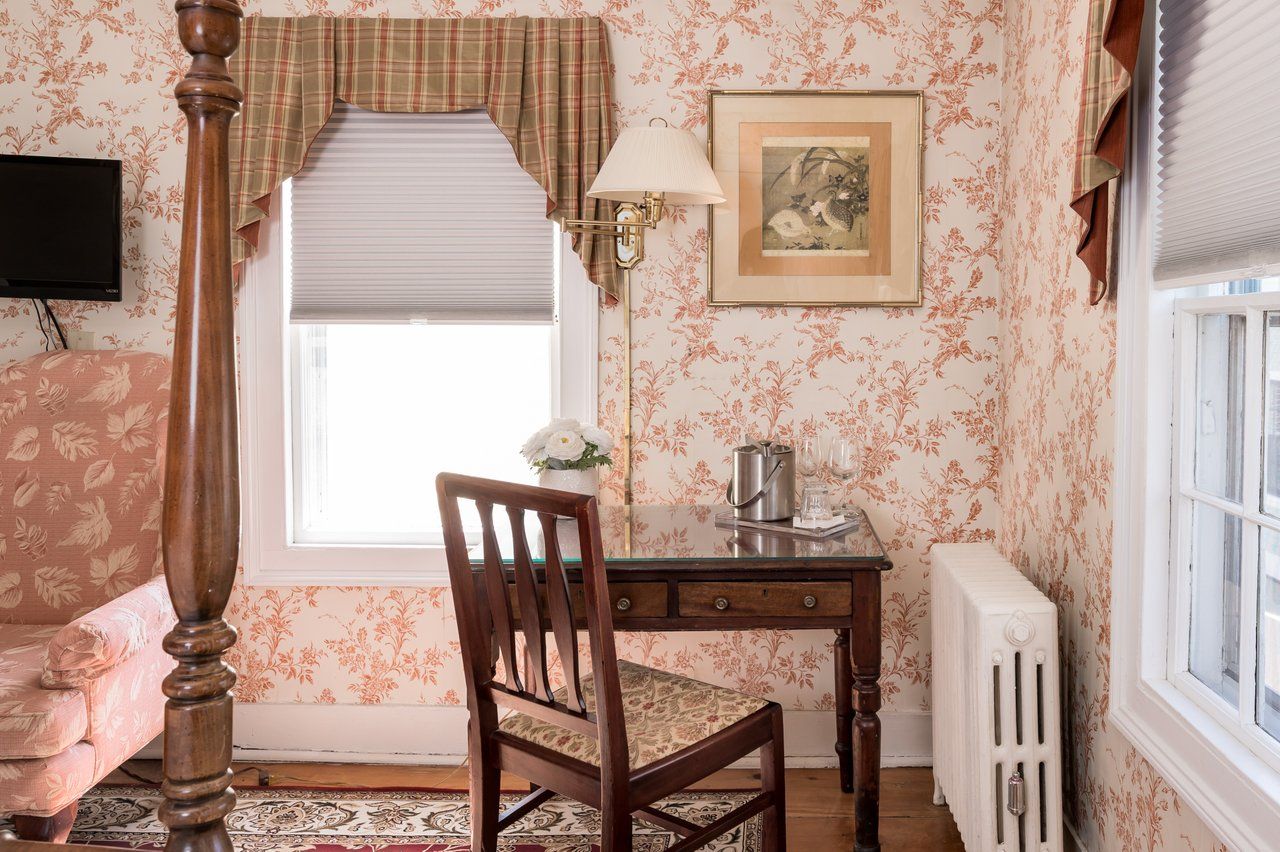A desk and chair sit in a bedroom corner with floral wallpaper, a wood-framed window, and a white radiator.