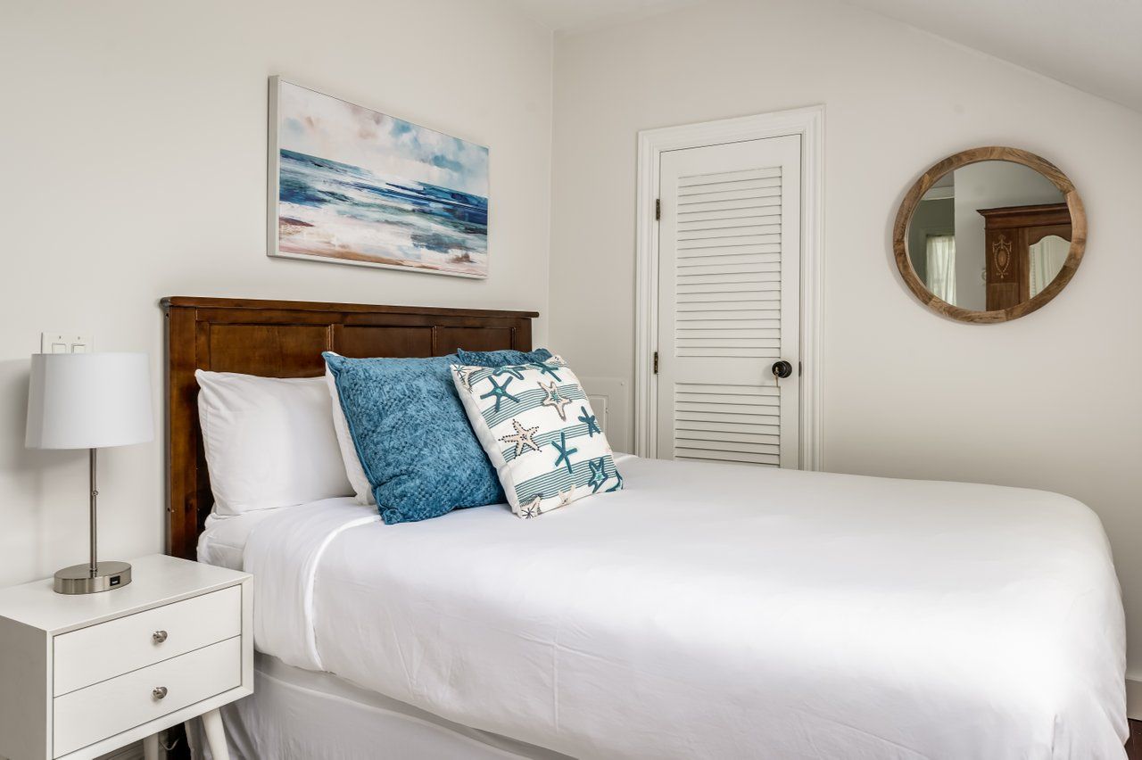 A white-walled bedroom with a queen bed, blue and white nautical-themed pillows, a white nightstand, and a round mirror.